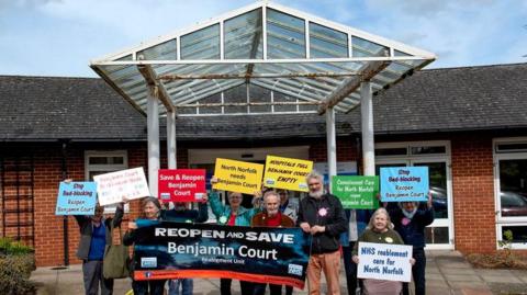 A brick building with a glass entrance with a group of protestors gathered outside holding banners reading "Save Benjamin Court"