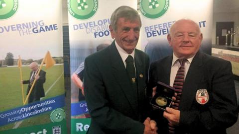 Two men wearing dark blazers and ties shake hands at the presentation of an award. One is holding a medal housed in a small black box. Behind them are colourful banners advertising the work of the Lincolnshire FA, including photos of footballers and officials and slogans such as "governing the game" and "growing opportunities across Lincolnshire".