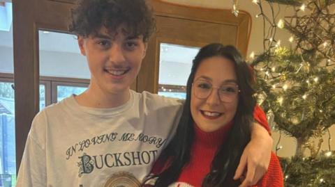 Corrin Wallace and her son Joshua are standing inside a house in front of windowed double-doors. There is a Christmas tree and lights in the corner of the photo. Corrin is wearing a red jumper and glasses. Joshua is wearing a white T-shirt with the words "Buckshot" on it. They are standing next to each other and he has his left arm over her left shoulder.