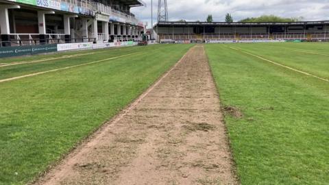 A green football pitch with two stands in the background - one facing the pitch on the right on the photo and the other on the left is at an angle to the camera. Although the pitch is mainly green, there is a strip that is soil going from the bottom of the image in the middle up to nearly the top of the photo. 