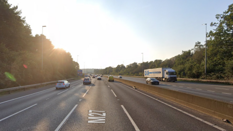 A Google street view screenshot of a four-lane motorway with cars and a lorry driving on it.