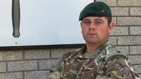 Danny Bowcock, who has his arms folded and is standing in front of a brick wall, is wearing a camouflage uniform and a dark green beret.