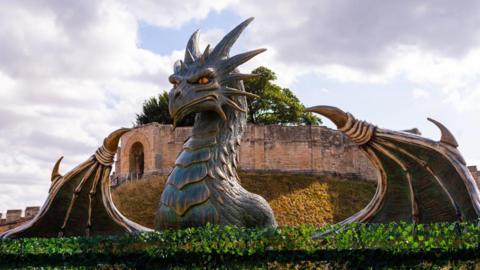 A large sculpture of a grey dragon with its wings outstretched is surveying the scene with striking yellow eyes. Castle walls are behind it.
