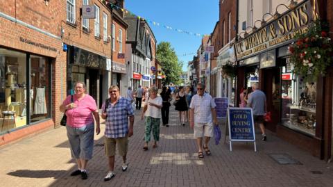 A pedestrianised street with a large number of people walking along it. There are shops either side, with hanging baskets and bunting also visible. Some of the shops have A-boards outside of them.