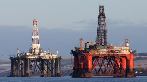 Two oil platforms in the North Sea. The one in the foreground is red and has a white cabin with a black tower on top. In the background, the platform is black with a white cabin and white and red tower.