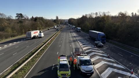 A picture of a motorway from a bridge above. The coastbound carriageway, on the left, is open with traffic flowing. The London-bound carriageway, on the right, is blocked off by Highways Officers and traffic is exiting via a slip road.