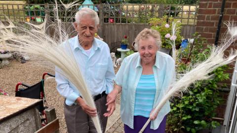 An elderly couple holding large white feathers from a peacock. The man on the left is wearing grey trousers and a blue shirt and has grey hair. The woman on the right is wearing a shirt over a blue top and is holding onto her husband's arm. They are in a garden with a fence and plants in the background.
