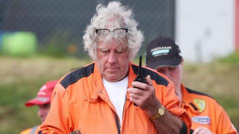 Maurice Bennington, wearing a white T-shirt, orange jumpsuit, holding a walkie-talkie in his left hand, with a gold watch on. He is grey hair and there are two people behind him also wearing orange jumpuits and wearing caps.