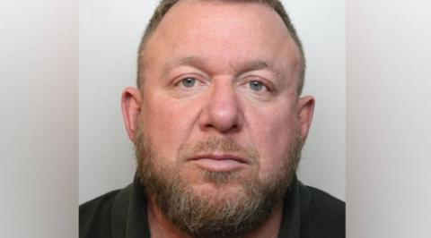 A police custody head and shoulders mugshot of Mark Killick. He has a scruffy dark brown beard and receding hair on his head and is wearing a dark coloured polo top