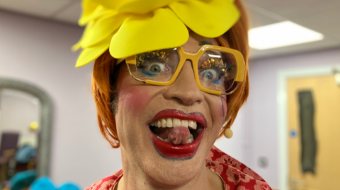 A man in thick make up, wearing big yellow framed glasses and a ginger wig is sticking his tongue out at the camera