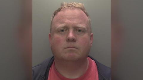 Robert Bush with short, light brown hair is shown in a straight‑on mugshot photograph. He has a neutral expression and is wearing a dark jacket over a red shirt. The background is plain and grey, and a Humberside Police badge watermark appears in the upper right corner of the image.
