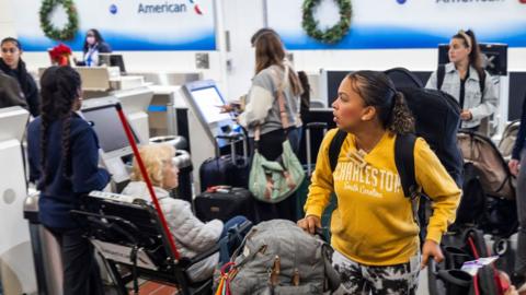 A woman in a yellow top with luggage waits with other people at a check in desk at an airport. She is carrying a backpack and looking to the left