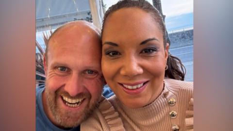 Stock and Caz are pictured smiling at the camera in a selfie. She has her hair tied back and a beige ribbed jumper with frilled shoulders and gold buttons. He is wearing a blue tee shirt and has a bald head and ginger beard.