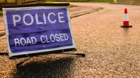 A stock image of a roundabout at dawn with a police road closed sign and orange traffic cones.