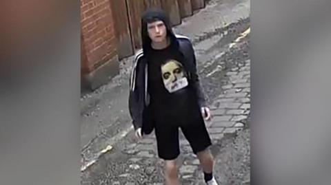 CCTV image of a man with black hair wearing an unzipped, hooded black jacket with white stripes running halfway down each sleeve. He is also wearing a black t-shirt with a man's grey face on it, and black socks and white trainers. He is walking down an alley. 
