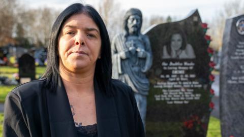 Helen looks straight at the camera. She wears a black suit jacket and has black hair. She stands in a graveyard in front of her daughter Demi's headstone.