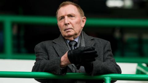 Sir Tom Farmer standing in a football stand. he is wearing a greay coat and grey shirt and tie