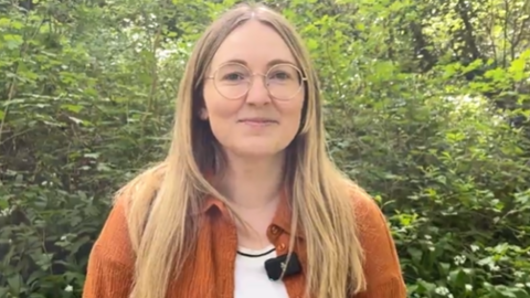 Becca is standing in front of a green hedge in the sun. She is wearing an orange cardigan and has long, blonde hair and glasses.