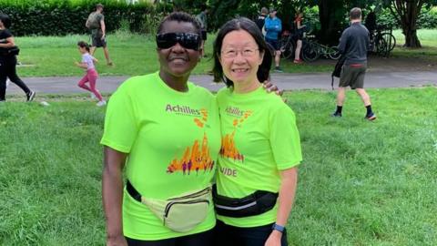 Judith Namaganda, wearing a bright green top and black sunglasses, with her arm around another runner who is wearing a bright green top, has glasses and shoulder-length dark hair