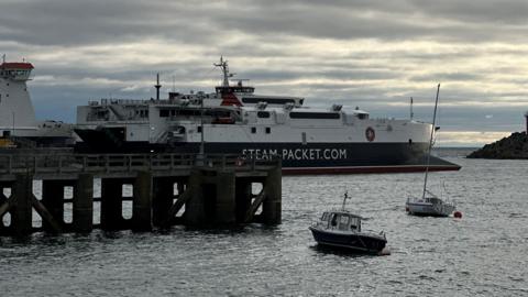 The Manannan, a sleek catamaran which is red, white and black and has Steam-Packet.com on the side in white lettering, as it leaves Douglas Harbour.