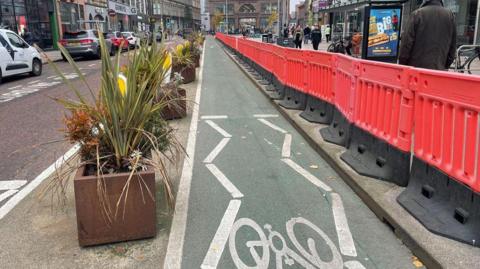 A cycle lane which in lined with an orange barrier on the kerb. People can be seen walking on the footpath past some shops. 