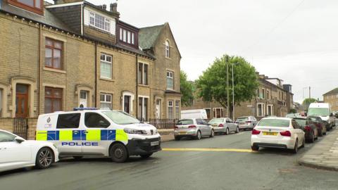 A police van is parked across a street in Bradford which has terraced houses on each side.