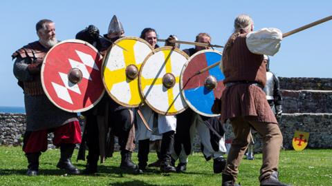 A colourful image of knights dressed in traditional costume, with their armoury, re-enacting a scene from medieval times, amidst the grounds of Castle Cornet and blue skies overhead.