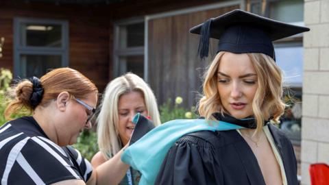 A woman with curled blonde shoulder length hair is looking towards the ground and wearing a light yellow dress and gold necklace underneath a black graduation gown and cap. The back of the gown is blue and two women are smiling as they sort it out for her. You can see a brown building and shrubbery in the background.