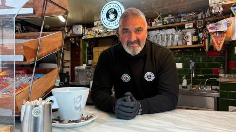 Liberato Lionetti with short white hair and beard, wearing a black top with two circular logos on it, and grey gloves, stands behind the counter of a coffee shop. There are packs of coffee behind him on wooden shelves, a wooden display shelf to his right and a grey coffee cup in the foreground