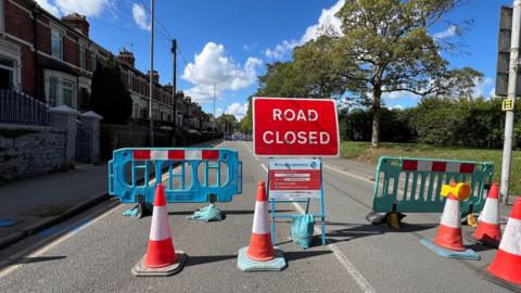 Road closed sign, barriers and cones in a road, with red brick terraced homes to the left and greenery on the right. A sunny day.