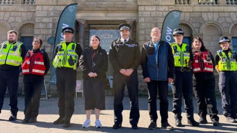 Nine people stand in a line, some police officers wearing yellow high vis vests, and some city officers wearing red vests.