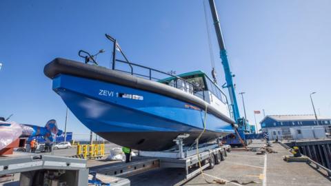 A bright blue electric boat called Zevi 1 sits on top of a wheeled metal trolley on dry land
