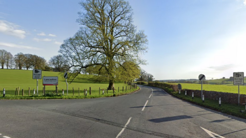 A sign says 'Lancashire' and two other signs say 'Average speed check'. There are trees and fields either side of the road.