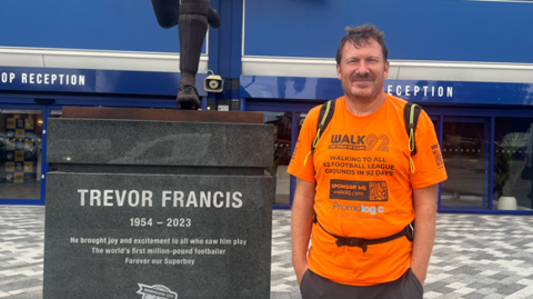 Xavier Wiggins is dressed on the right in bright orange top that says Walk 92. He's stood next to the Trevor Francis memorial bronze statue outside St.Andrews at Knighthead Park.