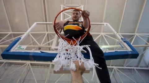 Ben sat on top of an indoor basketball hoop wearing a black vest.