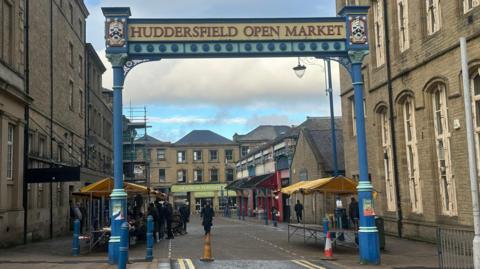 York stone buildings tower above iron pillars and a gateway sign that reads Huddersfield Open Market. Beyond the archway, traders under yellow canopies line streets.