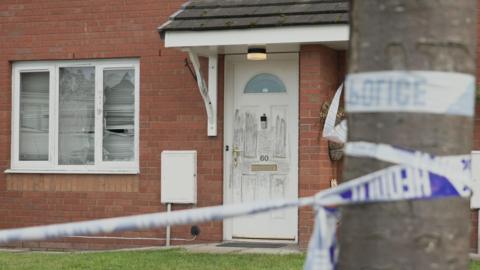 Police tape tied to a tree outside a red-brick house with a porch and a white door.