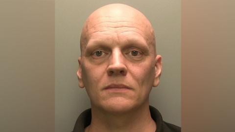 Ben Cairns has brown eyes and is clean shaven with no hair on his head.