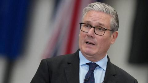 Sir Keir Starmer speaks, wearing spectacles, a dark suit, light blue shirt and a navy blue tie, against a blurred background