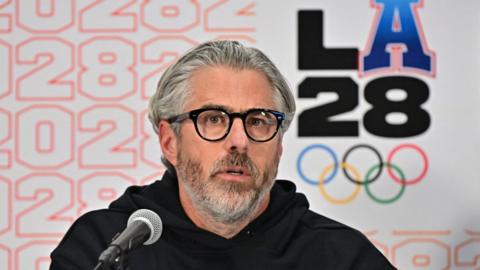 Casey Wasserman speaks to the media in Los Angeles in June 2025. He is wearing a black sweatshirt and glasses.
