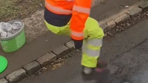 Legs in orange and yellow hi-vis are seen jumping on a newly-repaired pothole on a rural road