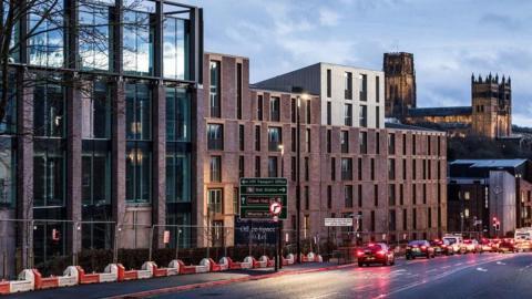 Milburngate. The development is several storeys high and has large glass windows. Durham Cathedral can be seen in the background and a number of vehicles are being driven along the main road in front of the development.