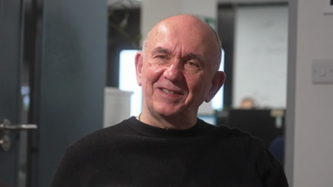 Peter Molyneux is sitting in his office looking off camera during an interview, wearing a black jumper