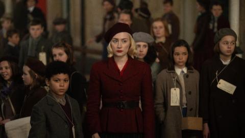 A still from the film Blitz, with a woman and a boy dressed in 1940s clothing. They are surrounded by children playing evacuees. The woman is wearing a red coat with a black belt around her waist and with a matching red beret. The children have what appears name tags around their neck.