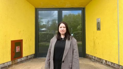 A woman with dark hair and a check coat stands outside a building with yellow walls and a glass door.