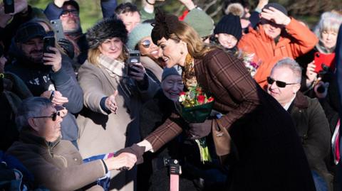 Princess of Wales holding a bouquet of flowers in her left had and she shakes hands with an elderly man sitting down. Around them are well-wishers