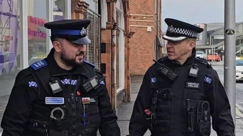 Two officers - a Police Community Support officer on the left in black uniform with blue detailing - and Chief Superintendent Owain Llewellyn on the right in a black uniform with white detailing.  Both are on the beat in Water Street in Rhyl town centre.