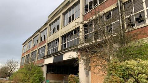 A former car factory with smashed windows and trees growing around it on a grey day