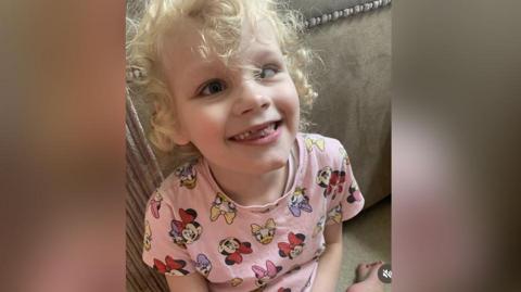 Seven-year-old Isla with blond curly hair wearing a pink Disney patterned t-shirt sits on the floor leaning on a beige couch. She is smiling.