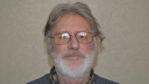 A man with grey hair that has grown down to his ears. He also has a thick grey beard. He is wearing glasses along with a grey jumper and chequered green shirt.